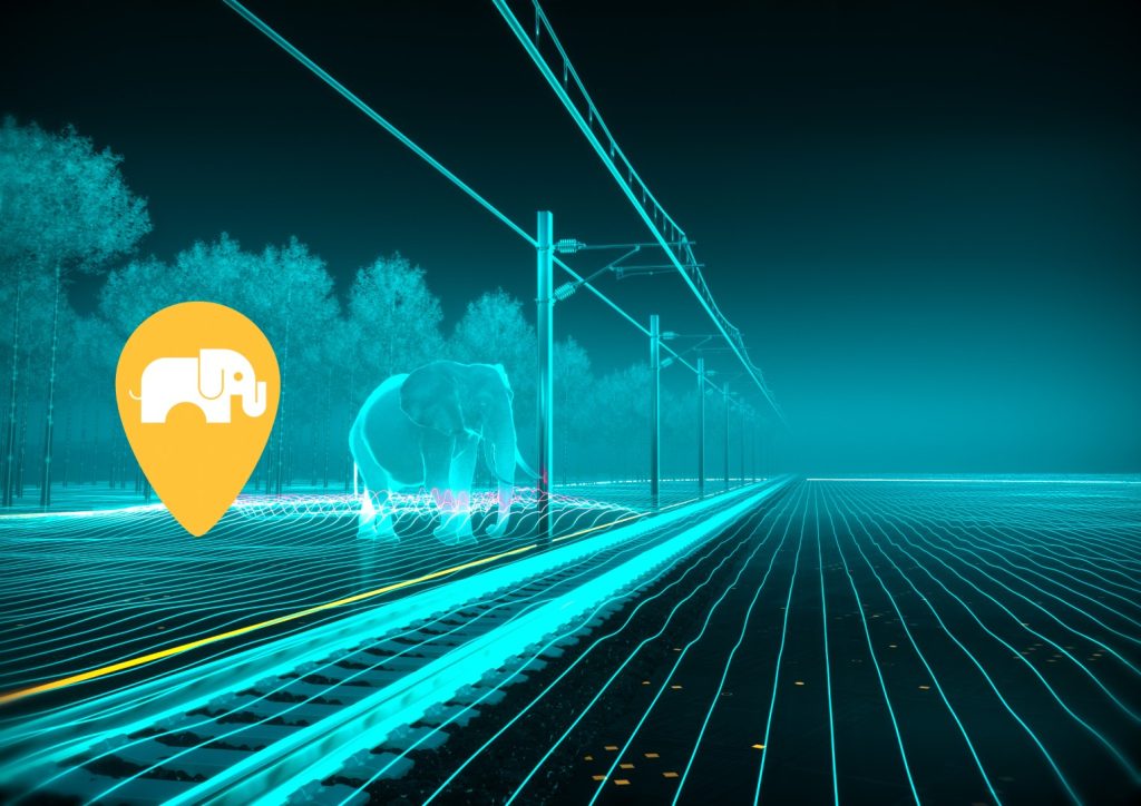 A computer-generated image of an elephant walking across a railway with a forest in the background. Glowing orange fiber optic cable runs alongside the track. A large warning sign/pin of an elephant is next to the elephant.